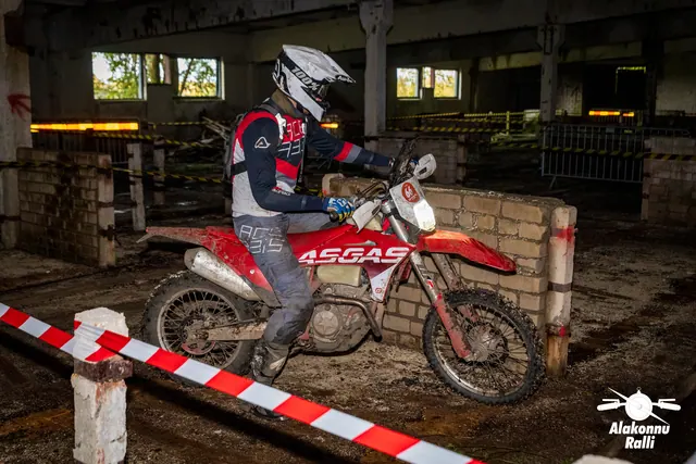 Alakonnu Ralli 2026: valge kiivriga sõitja juhib punast mootorratast hoones punavalge rajalindi vahel.