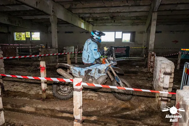 Alakonnu Ralli 2026: helesinises sõiduvarustuses osaleja juhib mootorratast hoones punavalge rajalindi kõrval.