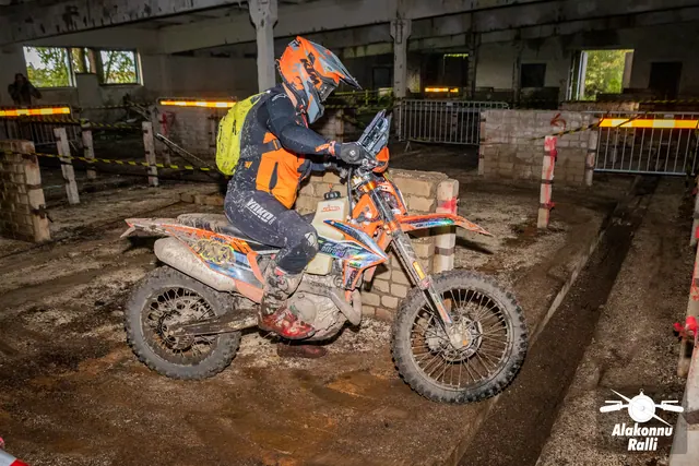 Alakonnu Ralli 2026: oranži kiivri ja kollase seljakotiga sõitja juhib oranži mootorratast hoones rajalintide vahel.