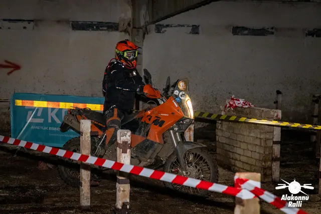 Alakonnu Ralli 2026: oranži kiivriga sõitja juhib oranži mootorratast hoones tellismüüri ja rajalindi vahel.