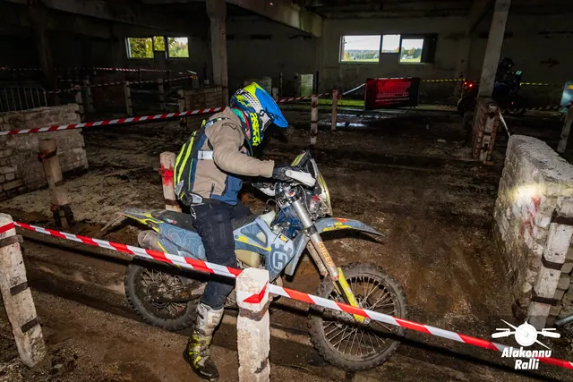 Alakonnu Ralli 2026: sinise ja kollase kiivriga sõitja juhib valget mootorratast hoones punavalge rajalindi kõrval.