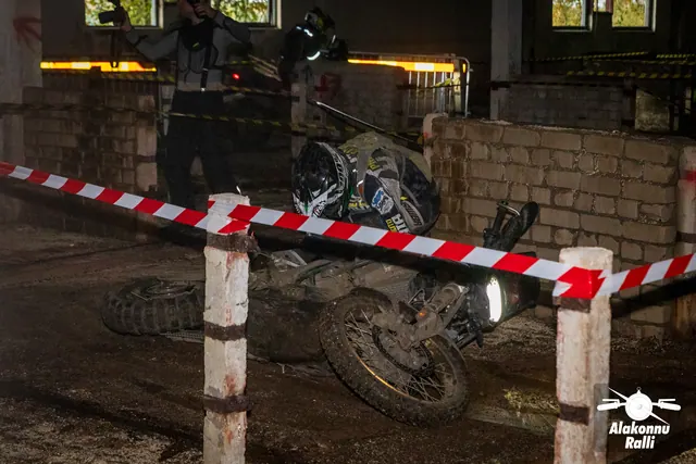 Alakonnu Ralli 2026: kiivriga sõitja kummardub hoones rajalintide vahel külili kukkunud mootorratta kohale.