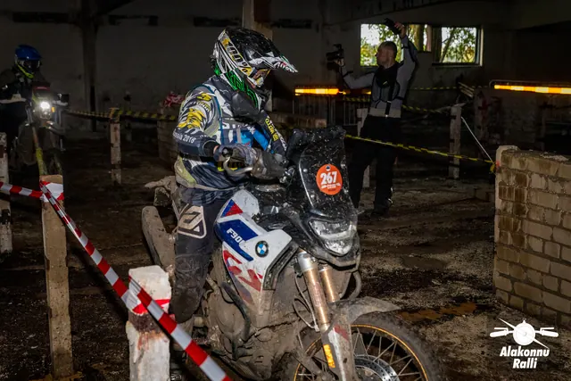 Alakonnu Ralli 2026: mustrilise sõiduvarustusega osaleja juhib heledat mootorratast hoones punavalge rajalindi vahel.