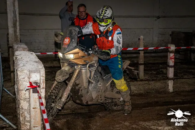 Alakonnu Ralli 2026: osaleja juhib porist mootorratast hoones madala müüri kõrval, oranži logoga pusas korraldaja seisab taga.