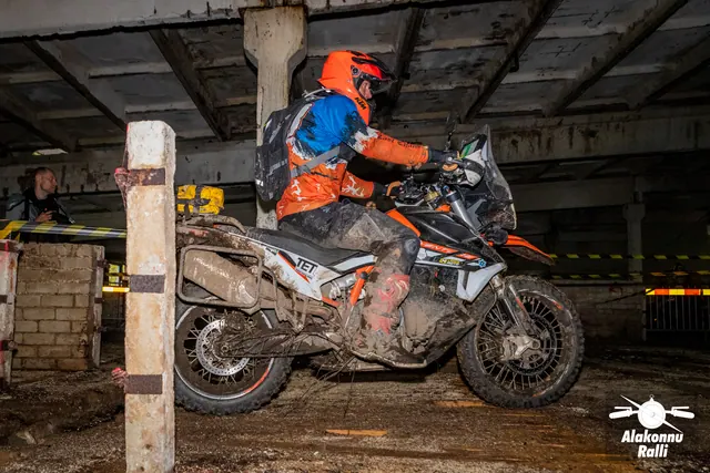 Alakonnu Ralli 2026: oranži kiivriga sõitja juhib porist mootorratast hoones punavalge rajalindi kõrval.