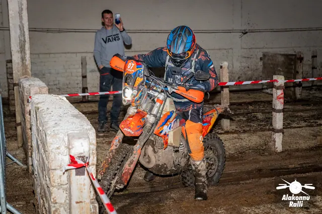 Alakonnu Ralli 2026: oranžis ja sinises sõiduvarustuses osaleja juhib oranži mootorratast hoones, taustal inimene telefoniga.