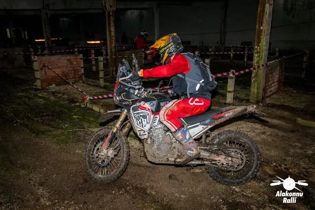 Alakonnu Ralli 2026: punase sõiduvarustusega osaleja juhib porist mootorratast hoones rajalintide vahel.