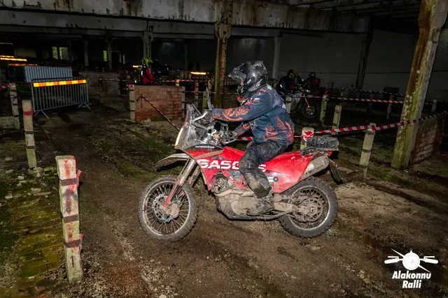 Alakonnu Ralli 2026: tumedas sõiduvarustuses osaleja juhib punast mootorratast hoones rajalindi kõrval.