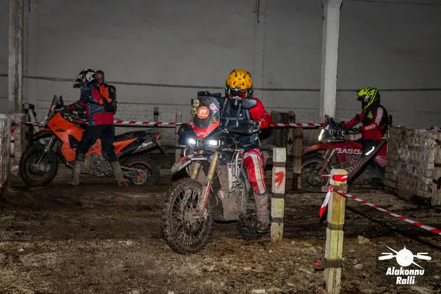 Alakonnu Ralli 2026: kolm mootorratturit koguneb hoones rajalintidega piiratud radade ristumiskoha juurde.