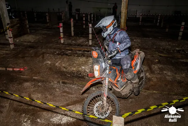 Alakonnu Ralli 2026: valge kiivriga sõitja juhib oranži mootorratast hoones kollase ja musta rajalindi taga.