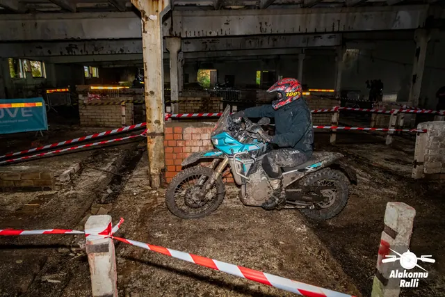 Alakonnu Ralli 2026: tumedas sõiduvarustuses osaleja juhib sinist mootorratast hoones punavalge rajalindi kõrval.