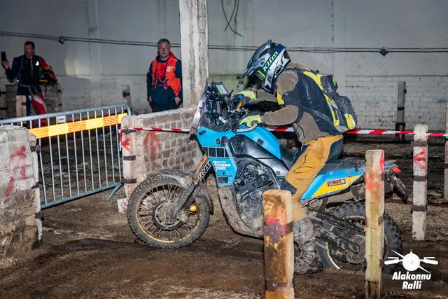 Alakonnu Ralli 2026: sõitja juhib sinist mootorratast hoones metallpiirde ja rajalindi vahel; taustal jälgib Alakonnu logoga oranžis pusas korraldaja.
