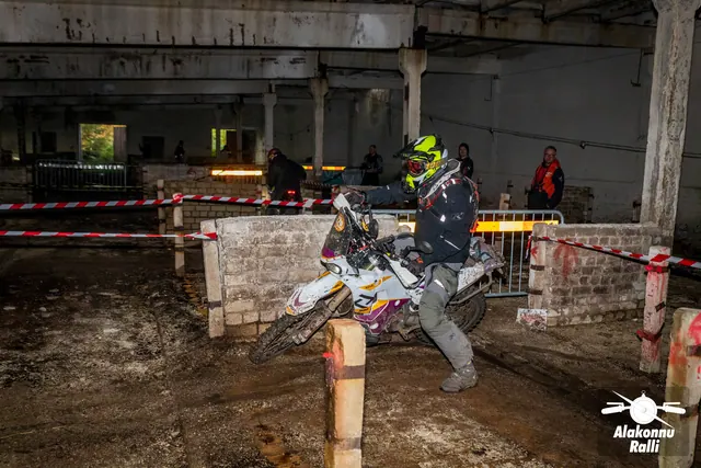 Alakonnu Ralli 2026: sõitja pöörab valge mootorrattaga hoones madala tellismüüri kõrval; taustal seisab Alakonnu logoga oranžis pusas korraldaja.