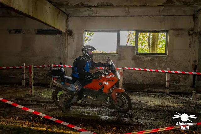 Alakonnu Ralli 2026: mootorrattur juhib oranži mootorratast hoones rajalintide vahel, akendest paistab päevavalgus.
