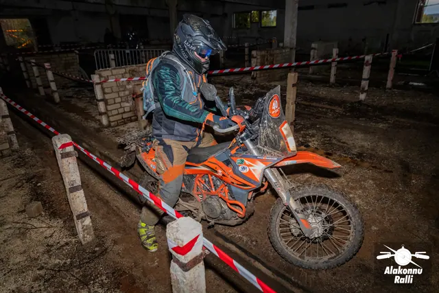 Alakonnu Ralli 2026: roheliste varrukatega sõitja juhib oranži mootorratast hoones punavalge rajalindi vahel.