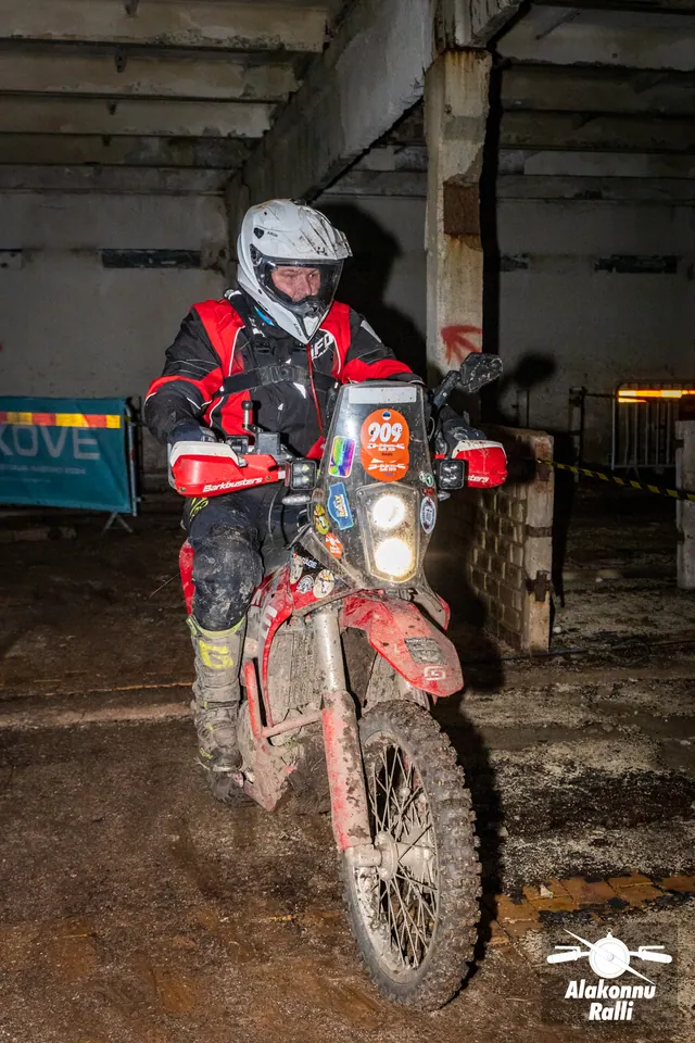 Alakonnu Ralli 2026: valge kiivriga ja punases sõiduvarustuses osaleja läheneb hoones porisel mootorrattal.