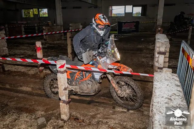 Alakonnu Ralli 2026: oranži kiivriga osaleja juhib oranži mootorratast hoones punavalge rajalindi kõrval.