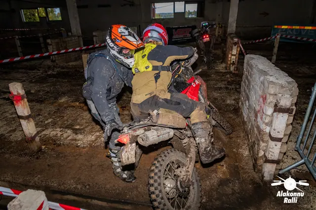 Alakonnu Ralli 2026: oranži kiivriga osaleja aitab punasel mootorrattal sõitjat hoones madala müüri kõrval.