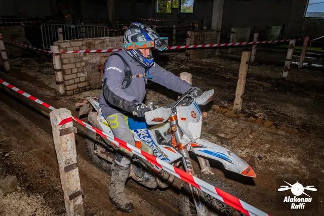 Alakonnu Ralli 2026: hallis ja sinises sõiduvarustuses osaleja juhib valget ja oranži mootorratast hoones.