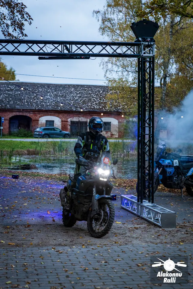 Alakonnu Ralli 2026: mootorrattur sõidab põleva esitulega läbi õue metallraami, ümber sinakas valgus ja suits.