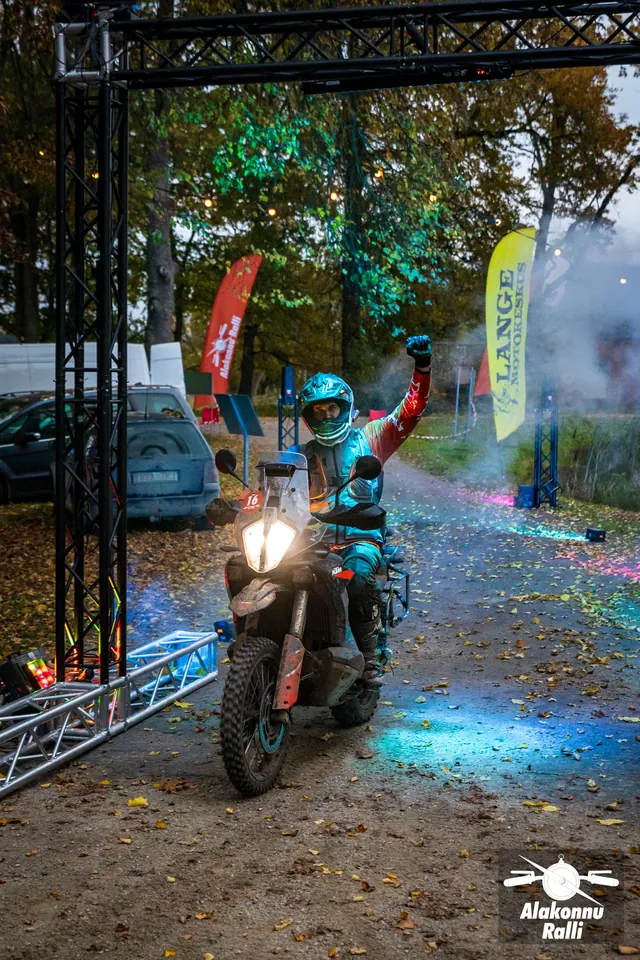 Alakonnu Ralli 2026: mootorrattur tõstab käe õues metallraami juures, ümber värviline valgus ja suits.