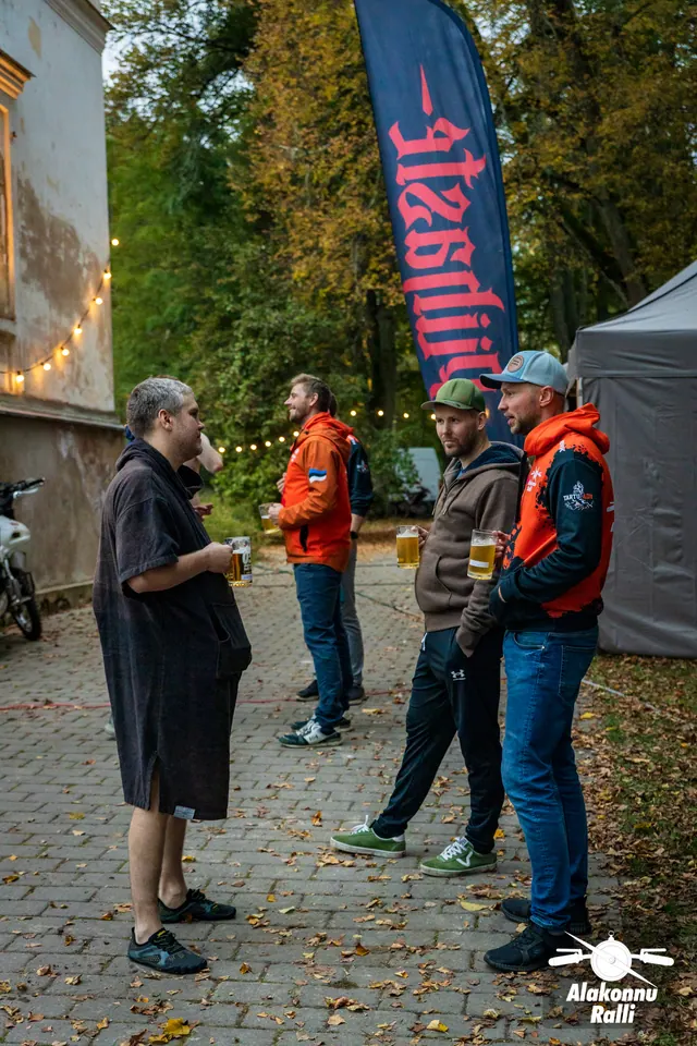 Alakonnu Ralli 2026: Alakonnu logoga oranžis pusas korraldaja ja teised rallikülalised vestlevad õues joogikruusidega.